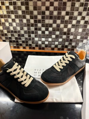 Maison Margiela svarta sneakers - Stilrena svarta sneakers från Maison Margiela med beige snörning och gummisula. Skorna har en kombination av mocka och skinn, samt klassisk låg profil och rund tå. Perfekta för dig som gillar minimalistisk och tidlös design.