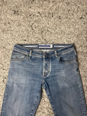 Ljusblå jeans från Jacob Cohen - Snygga ljusblå jeans från Jacob Cohen med klassisk femficksdesign och diskreta slitningar framtill. Det finns två defekter den ena är att bak patchen är väldigt skadad och den andra så finns det två slitningar som går igenom.
