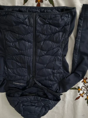 Svart cardigan  Peak Performance - Snygg svart cardigan från Peak Performance med quiltat mönster på framsidan, huva och dragkedja. Jackan har släta ärmar och två sidofickor med dragkedja. Perfekt för blåsiga dagar och outdoor-aktiviteter.