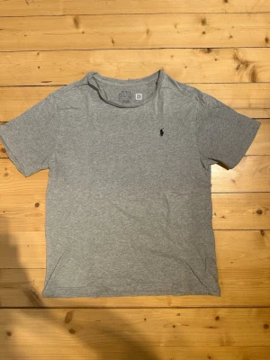 Grå t-shirt från Polo Ralph Lauren - Klassisk grå t-shirt från Polo Ralph Lauren med korta ärmar och rund hals. Liten mörkblå logga broderad på bröstet. Tillverkad i mjuk bomull för en skön och avslappnad stil. Tveka inte att skriva vid frågor eller funderingar 