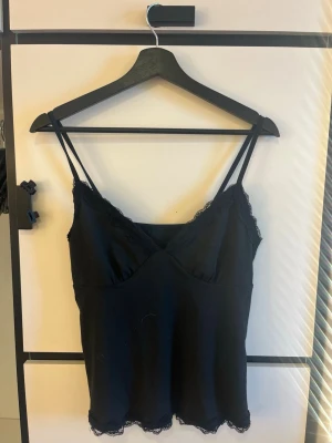 Svart spetsdetaljerad linnetopp - Snygg svart linnetopp med tunna axelband och spetsdetaljer längs v-ringningen och nederkanten. Perfekt att styla lager på lager eller som den är. Mjuk och bekväm känsla med en feminin touch.