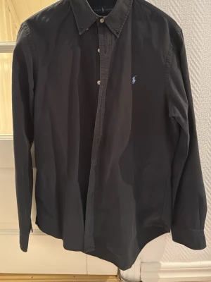 Mörkblå skjorta från Ralph Lauren - Klassisk mörkblå skjorta från Ralph Lauren med button-down krage och broderad logga på bröstet. Skjortan har långa ärmar och knäppning framtill. Perfekt till jeans eller chinos för en clean look.