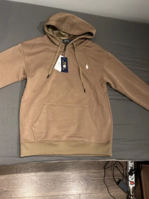 Beige hoodie från Polo Ralph Lauren - Snygg beige hoodie från Polo Ralph Lauren med klassisk huva och dragsnören. Har en känguruficka framtill och den ikoniska vita Polo-loggan broderad på bröstet. Tillverkad i mjukt bomullsmaterial för skön känsla. Perfekt för en avslappnad och stilren look.