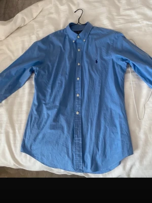 Blå skjorta från Ralph Lauren - Klassisk blå skjorta från Ralph Lauren i slim fit-modell. Skjortan har knappar framtill, button down-krage och det ikoniska broderade logotypen på bröstet. Tillverkad i bomull och har långa ärmar. Perfekt för en stilren look.