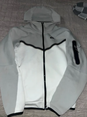 Nike vit och grå hoodie med dragkedja - Nike hoodie i vitt och ljusgrått med hel dragkedja framtill och svart detalj längs bröstet. Huva och en ficka med dragkedja på ärmen. Tillverkad i mjukt material, perfekt för chill eller träning. Klassisk Nike-logga på bröstet. Pris kan diskuteras storlek i annons stämmer inte skriv t mig för info