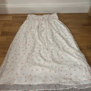 Vit blommig långkjol med snörning - Söt vit långkjol med små rosa blommor och smockad midja. Kjolen har snörning med tofsar i midjan och är gjord i ett lätt, luftigt tyg som passar perfekt till varmare dagar. Romantisk och flowy stil.