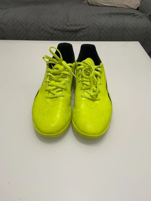Gula fotbollsskor från Puma - Snygga fotbollsskor från Puma i en skarp gul färg med svarta och vita detaljer. Skorna har snörning och låg profil, perfekt för snabba rörelser på planen. Ovandelen är i syntetmaterial och sulan är platt, vilket ger bra grepp på inomhusunderlag.