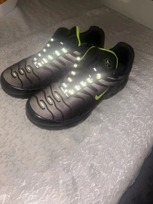 Nike sneakers med reflexdetaljer - Snygga Nike sneakers i svart och grått med neongröna detaljer och reflekterande snörning. Skorna har en sportig design med vågformade linjer på ovandelen och klassisk Nike-logga på sidan. Perfekta för dig som gillar streetwear och vill sticka ut.