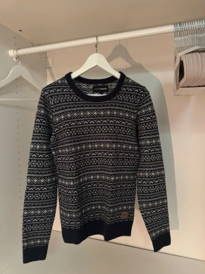 Marinblå & vit stickad tröja KappAhl - Snygg marinblå stickad tröja från KappAhl med klassiskt mönster i vitt. Tröjan har rund halsringning, långa ärmar och ribbade muddar vid ärmslut och nederkant. Perfekt för kalla dagar och ger en stilren look.