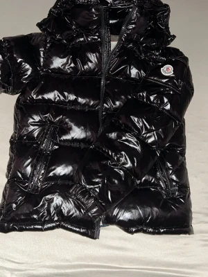 Svart Moncler maya  - Snygg svart dunjacka från Moncler med glansig finish och klassisk pufferstil. Jackan har huva, dragkedja framtill och två dragkedjefickor. Moncler-logga på ärmen ger en exklusiv touch. Jackan kommer med NFC (tar dig till webbsidan när du håller mobilen mot loggan)