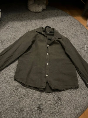 Olivgrön linneskjorta med knappar - Stilren olivgrön skjorta i linne med klassisk krage och vita knappar framtill. Skjortan har långa ärmar med knapp vid ärmslut och en avslappnad passform. Perfekt för dig som gillar en enkel och tidlös look.