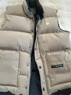 Beige/brun dunväst från Canada Goose - Stilren beige dunväst från Canada Goose med hög krage och två stora fickor framtill. Västen har dragkedja och tryckknappar samt diskret logga på bröstet och etikett på fickan. Normal i storlek 