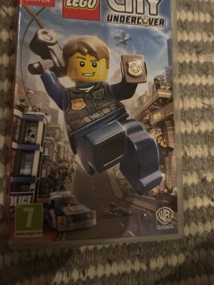 LEGO City Undercover till Switch - LEGO City Undercover är ett actionäventyrsspel till Nintendo Switch där du spelar som polis i en öppen värld byggd av LEGO. Omslaget är färgglatt med en LEGO-polisfigur i blå uniform och stadsmiljö i bakgrunden. Perfekt för dig som gillar LEGO och äventyr.