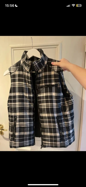 Rutig dunväst från Lauren Ralph Lauren - Snygg rutig dunväst från Lauren Ralph Lauren i svart, vitt och grått. Västen har hög krage, dragkedja framtill och två fickor med dragkedja. Tillverkad i mjukt material med quiltad look och klassiskt rutmönster. Perfekt för lager på lager under kyliga dagar.