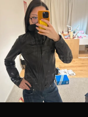 Svart y2k skinnjacka med dragkedja - Snygg svart skinnjacka med hög krage och dragkedja framtill. Jackan har en figurnära passform och diskreta sömdetaljer vid axlar och bröst. Perfekt för dig som vill ha en stilren och tidlös look.