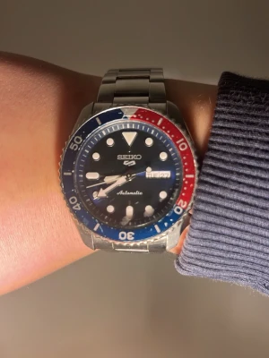 Seiko SRPD53 5 sports automatisk klocka Pepsi - Snygg Seiko 5 automatisk klocka med blå och röd vridring, blå urtavla och dag/datumvisning. Boett och länk i rostfritt stål. Genomskinlig baksida som visar urverket. Perfekt för dig som gillar sportig stil.                      Lite repor på ur tavlan.