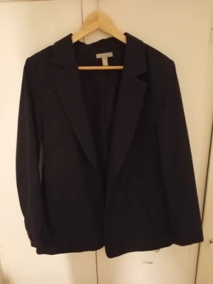Svart kavaj från H&M - Stilren svart kavaj från H&M med klassisk krage och två framfickor. Perfekt för en dressad look och enkel att matcha med olika outfits. Långärmad modell med normal passform.