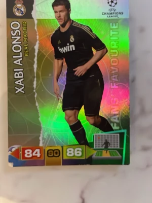 Xabi Alonso Real Madrid samlarkort - Samlarkort med Xabi Alonso från Real Madrid, Champions League-serien. Kortet har holografisk yta med färgglada effekter och visar spelaren i svart matchställ. Siffror och statistik syns på framsidan. Perfekt för fotbollsfans och samlare.
