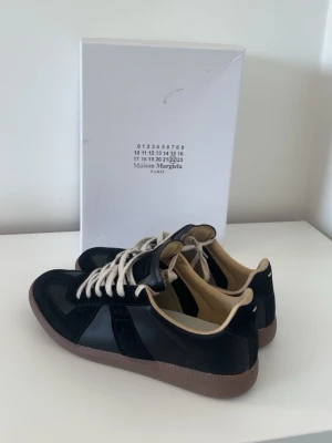 Maison Margiela svarta sneakers - Maison Margiela sneakers i svart mocka och skinn med beige insida och vit snörning. Sulan är i gummi med klassiskt mönster och rund tå. Snygg och stilren design som passar till många olika outfits.
