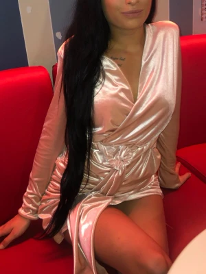 Champagnefärgad omlottklänning i satin - Snygg kort omlottklänning i champagnefärgad satin med lång ärm och knyt i midjan. Klänningen har en djup v-ringning och slits framtill för en trendig look. Perfekt för dig som vill sticka ut med en elegant och glansig stil.