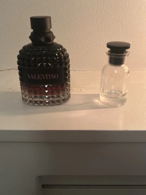 Valentino parfymflaska born in Roma intense  - Säljer en snygg parfymflaska från Valentino med svart lock och coolt nitat mönster i glaset. Flaskan har en mörk tonad botten och en svart etikett med Valentino-loggan. Perfekt för dig som gillar exklusiva dofter och stilren design.