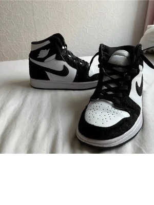 Nike Jordan 1 svartvita sneakers - Svartvita Nike Jordan 1 sneakers med klassisk high-top design. Skorna har svart swoosh, snörning och perforerad tåbox. Tillverkade i skinn och syntetmaterial för en stilren och sportig look.