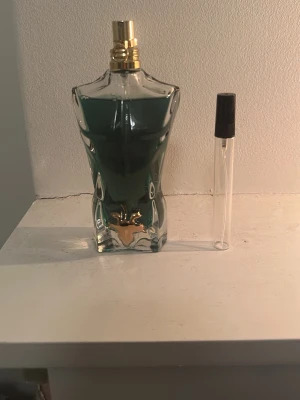 Le beau parfym från Jean Paul Gaultier - Säljer en Le beau parfym från Jean Paul Gaultier. Flaskan är formad som en manlig torso i genomskinligt glas med grönblå vätska och gulddetaljer vid halsen. En mindre provflaska med svart kork ingår.