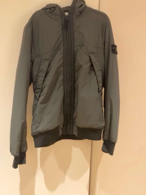Grön vindjacka från Stone Island - Snygg grön vindjacka från Stone Island med huva och dragkedja framtill. Jackan har ribbade muddar vid ärmar och midja samt den klassiska Stone Island-loggan på vänster ärm. Tillverkad i lätt, vindtätt material som passar perfekt till streetwear. Skriv vid intresse. Pris kan diskuteras.