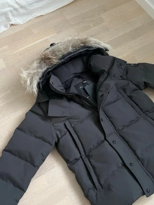 Grå dunjacka från Canada Goose - Säljer en svart dunjacka från Canada Goose med stor huva och avtagbar pälskant. Jackan har quiltad design, dragkedja och knappar framtill samt lång ärm. Perfekt för kalla vinterdagar och riktigt snygg streetstil.