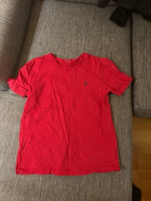 Röd t-shirt från Ralph Lauren - Klassisk röd t-shirt från Ralph Lauren med blå broderad logga på bröstet. T-shirten har rund halsringning och korta ärmar. Tillverkad i mjuk bomull som känns skön mot huden. Perfekt basplagg för en enkel och stilren look.