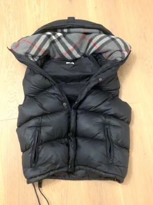 burberry väst  - säljer denna burberry väst med tanke på att ja inte använder den. Västen kan göras till en jacka om man har armarna till den vilket ja inte har. västen har ett hål inne i den men inget som skadar inget dun går ut från den. finns ett pytte litet hål i luvan men inget som märks eller gör någon skada. själva saken som man kan hänga västen med är sönder.