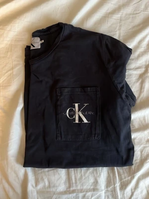 Svart t-shirt från Calvin Klein - Snygg svart t-shirt från Calvin Klein Jeans med stor ficka på bröstet och ikonisk CK-logga i vitt. Klassisk rund hals och korta ärmar. Perfekt till jeans eller shorts för en clean och stilren look.