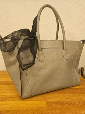 Grå rymlig tidslös handväska WERA Stockholm med svart scarf - Stilren grå handväska från WERA Stockholm Sparsamt använd, i gott skick. Passar för dig som vill rymma din laptop samt din mobil och kanske en liten necessär. (scarf ingår ej)