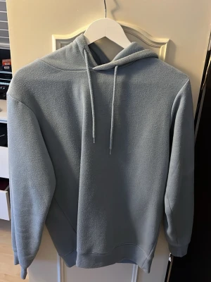 Ljusblå hoodie från SMOG - Snygg ljusblå hoodie från SMOG i regular fit. Hoodien har en klassisk huva med dragsko och är tillverkad i ett mjukt, fleeceliknande material. Perfekt för en avslappnad och stilren look.