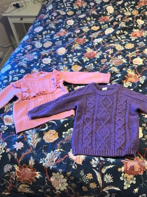 Lila och rosa stickade tröjor - Två stickade tröjor, en i lila med klassiskt flätmönster och ribbade muddar, samt en i rosa med volangdetaljer och strukturstickning. Båda har rund hals och långa ärmar. Perfekta för dig som gillar färg och mysiga plagg. Back finns 2 knappar för att lättare ta på tröjan