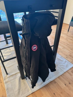 Svart jacka från Canada Goose - Säljer en svart jacka från Canada Goose med klassisk logotyp på ärmen. Jackan har huva, dragkedja framtill och flera praktiska fickor. Perfekt för kalla dagar och riktigt snygg till vardagslooken. Inga skador äkta jacka har använt i 2 månader.