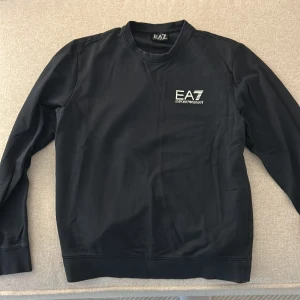 Svart EA7 långärmad sweatshirt - Svart långärmad sweatshirt från EA7 Emporio Armani med vit logotyp på bröstet och diskreta ränder i nacken på baksidan. Tröjan har rund hals och är tillverkad i mjuk bomull med en clean och stilren look. Perfekt för dig som gillar sportig och modern stil.