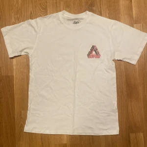 Vit Palace t-shirt med grafiskt tryck - Vit t-shirt från Palace med stort grafiskt Tri-Ferg tryck i rosa och svart på ryggen och mindre tryck på bröstet. Klassisk passform och rund hals. Materialet är mjuk bomull, perfekt för dig som gillar streetwear och snygga detaljer.