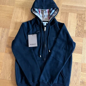 Svart/mörkblå burberry hoodie med dragkedja - Svart/mörkblå hoodie från Burberry med klassisk logga broderad på bröstet och ikoniskt rutigt foder i huvan. Tröjan har dragkedja framtill, snörning i huvan och ribbade muddar. Tillverkad i mjuk bomull för en skön känsla. Storlek M men passar bättre i S enligt mig