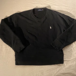 Svart sweatshirt från Polo Ralph Lauren - Svart sweatshirt från Polo Ralph Lauren med klassisk rund hals och broderad vit logga på bröstet. Tröjan har långa ärmar och ribbade muddar vid ärmslut och nederkant. Perfekt för en chill och stilren look.
