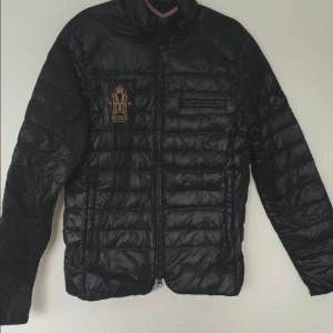 Morris svart jacka, storlek X-SMALL Unisex -   Går bra som XS till S  Morris Blake Light Jacka En svart jacka med dragkedja framtill och en broderad Morris-logga på vänster bröst. Jackan är lätt vadderad med en fyllning bestående av 90% dun och 10% fjäder, vilket ger utmärkt värme utan att kännas tung. Tillverkad i 100% nylon både på skal och foder, vilket gör den vind- och vattenavvisande samt be