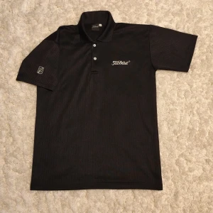 Svart Titleist piké i storlek L - Svart pikétröja från Titleist med diskret rutmönster och broderad logga på bröstet. Klassisk krage, två knappar och korta ärmar. Perfekt för golfbanan eller när du vill ha en sportig look.