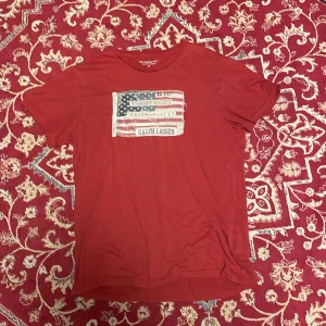 Röd t-shirt Ralph Lauren Denim & Supply - Röd t-shirt från Ralph Lauren Denim & Supply med coolt tryck av amerikansk flagga och broderade detaljer framtill. Klassisk rund hals och korta ärmar. Skön och mjuk bomullskvalitet, perfekt för en avslappnad stil.