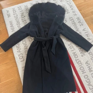 Svart kappa med fuskpäls och bälte - Stilren svart kappa med stor huva kantad av mjuk fuskpäls. Kappan har knytbälte i midjan och långa ärmar, perfekt för kalla dagar. Klassisk och elegant look som passar till många outfits.