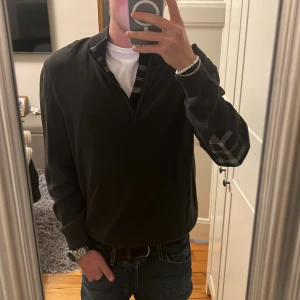 Grå Barbour half zip tröja M - Grå långärmad tröja från Barbour med half zip och rutiga detaljer på armbågar och insida krage. Klassisk och stilren modell i mjukt material, perfekt för lager-på-lager. Ribbad krage och muddar för extra komfort.