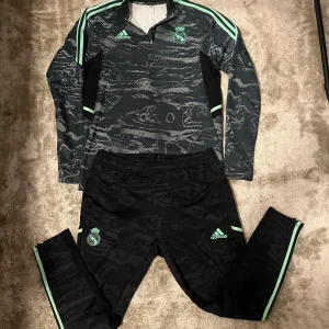Real Madrid Adidas träningströja M - Säljer en svart och mintgrön långärmad träningströja från Adidas x Real Madrid. Tröjan har ett unikt mönster, halv dragkedja och klubbens logga på bröstet. Perfekt för fotbollsträning eller chill. Snygga detaljer med mintgröna ränder på ärmarna.