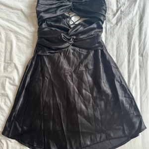 Svart satin klänning Selfie Leslie - Supersnygg svart kortklänning från Selfie Leslie i satinliknande material. Klänningen har snörning och cut out-detaljer framtill och baktill, samt en twistad detalj vid bysten. Perfekt för dig som vill sticka ut med en trendig look.