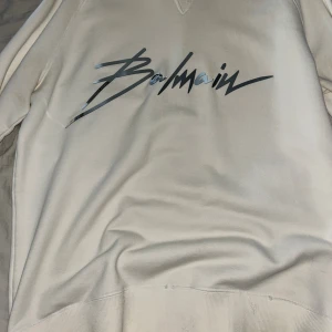 Vit sweatshirt från Balmain - Säljer en stilren vit sweatshirt från Balmain med stor logga r svart på bröstet. Tröjan har klassisk passform, rund halsringning och mjukt material som känns skönt mot huden. Perfekt för dig som gillar exklusiva streetwear-vibbar.