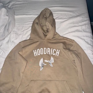 Beige hoodie från Hoodrich med tryck - Säljer en beige hoodie från Hoodrich med vit logga och två stilrena duvor tryckta på bröstet. Klassisk känguruficka och huva. Perfekt för dig som gillar streetwear och vill ha en snygg och bekväm tröja.