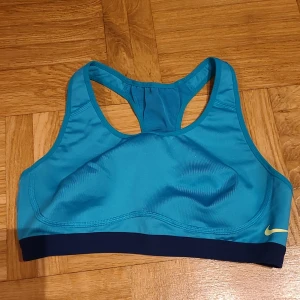 Blå sporttopp från Nike Dri-FIT - Sportig blå linnetopp från Nike med Dri-FIT-teknologi. Modellen har brottarrygg och bred mörkblå resår nedtill samt en liten gul Nike-logga framtill. Perfekt för träning och aktiva dagar.